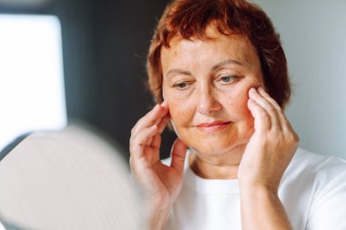 Küçük aynaya bakan ve rahat bir odada otururken yüzüne dokunan yetişkin kadın portresi. Kadın kırışıklıkları inceliyor, güneşli bir günde doğal güzelliğin tadını çıkarıyor. Yaşlanma kavramları, doğal güzellik..