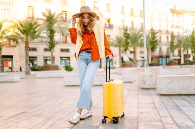 Şapka takan ve sarı bavul taşıyan kadın turist şehrin güneşli sokaklarında geziyor. Mutlu kadın küçük bavuluyla maceradan zevk alıyor ve yeni manzaralar keşfediyor. Turizm konsepti, hafta sonu.