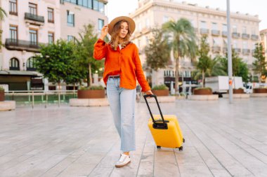 Şapka takan ve sarı bavul taşıyan kadın turist şehrin güneşli sokaklarında geziyor. Mutlu kadın küçük bavuluyla maceradan zevk alıyor ve yeni manzaralar keşfediyor. Turizm konsepti, hafta sonu.