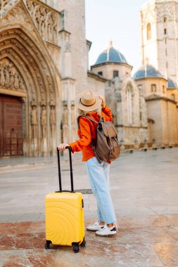 Şapka takan ve sarı bavul taşıyan kadın turist şehrin güneşli sokaklarında geziyor. Mutlu kadın küçük bavuluyla maceradan zevk alıyor ve yeni manzaralar keşfediyor. Turizm konsepti, hafta sonu.