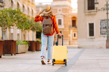 Şapka takan ve sarı bavul taşıyan kadın turist şehrin güneşli sokaklarında geziyor. Mutlu kadın küçük bavuluyla maceradan zevk alıyor ve yeni manzaralar keşfediyor. Turizm konsepti, hafta sonu.