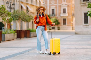 Şapka takan ve sarı bavul taşıyan kadın turist şehrin güneşli sokaklarında geziyor. Mutlu kadın küçük bavuluyla maceradan zevk alıyor ve yeni manzaralar keşfediyor. Turizm konsepti, hafta sonu.