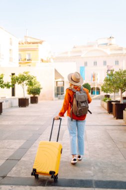 Şapka takan ve sarı bavul taşıyan kadın turist şehrin güneşli sokaklarında geziyor. Mutlu kadın küçük bavuluyla maceradan zevk alıyor ve yeni manzaralar keşfediyor. Turizm konsepti, hafta sonu.