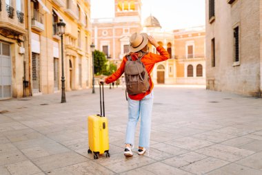 Şapka takan ve sarı bavul taşıyan kadın turist şehrin güneşli sokaklarında geziyor. Mutlu kadın küçük bavuluyla maceradan zevk alıyor ve yeni manzaralar keşfediyor. Turizm konsepti, hafta sonu.