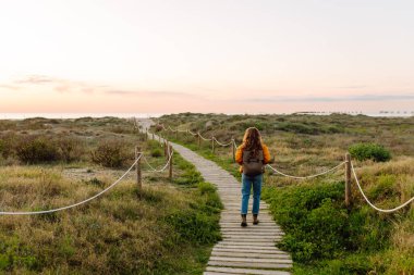 Sırt çantalı genç bir kadın şafak vakti plaja giden ahşap bir patika boyunca yürüyor. Parlak bir ceket giyen kadın deniz manzarasını ve yürüyüşü seviyor. Macera, yürüyüş konsepti.