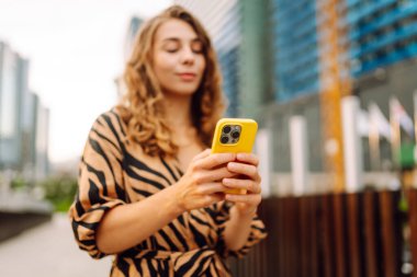 Güneşli bir günde şehir caddesinde telefonla dikilen genç bir kadının portresi. Elbise bloglarında bir turist ya da cep telefonunda Metropolitan City 'nin arka planına karşı mesajlar. Kavram: kentsel, blog yazarlığı, turizm.