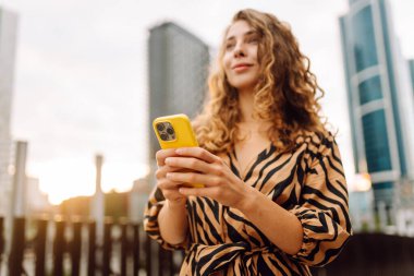 Güneşli bir günde şehir caddesinde telefonla dikilen genç bir kadının portresi. Elbise bloglarında bir turist ya da cep telefonunda Metropolitan City 'nin arka planına karşı mesajlar. Kavram: kentsel, blog yazarlığı, turizm.