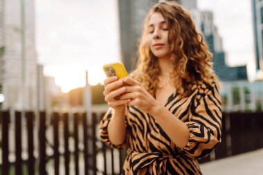 Güneşli bir günde şehir caddesinde telefonla dikilen genç bir kadının portresi. Elbise bloglarında bir turist ya da cep telefonunda Metropolitan City 'nin arka planına karşı mesajlar. Kavram: kentsel, blog yazarlığı, turizm.