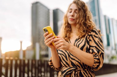 Güneşli bir günde şehir caddesinde telefonla dikilen genç bir kadının portresi. Elbise bloglarında bir turist ya da cep telefonunda Metropolitan City 'nin arka planına karşı mesajlar. Kavram: kentsel, blog yazarlığı, turizm.