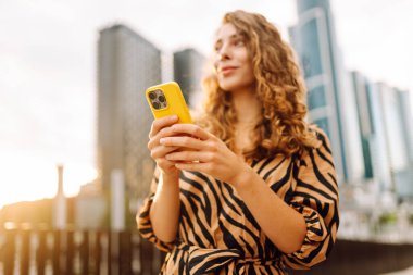 Güneşli bir günde şehir caddesinde telefonla dikilen genç bir kadının portresi. Elbise bloglarında bir turist ya da cep telefonunda Metropolitan City 'nin arka planına karşı mesajlar. Kavram: kentsel, blog yazarlığı, turizm.