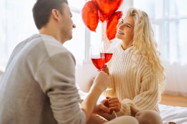 Genç çift, parlak bir odada bir kadeh şarapla birlikte oturur. Erkek, kız arkadaşına balonla süslenmiş bir odada hediyesini sunar. Randevularının tadını çıkarırlar. Tatil ve aşk konsepti. 14 Şubat.