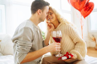 Genç çift, parlak bir odada bir kadeh şarapla birlikte oturur. Erkek, kız arkadaşına balonla süslenmiş bir odada hediyesini sunar. Randevularının tadını çıkarırlar. Tatil ve aşk konsepti. 14 Şubat.