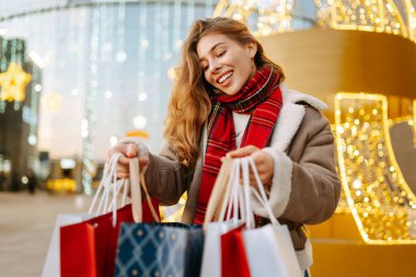 Alışveriş torbaları taşıyan neşeli bir kadın modern bir alışveriş merkezinin arka planında duruyor. Kadınlar açık havada alışveriş yaptıktan sonra şenlik havasına girerler. Konseptler: indirim, kutlama, alışveriş.