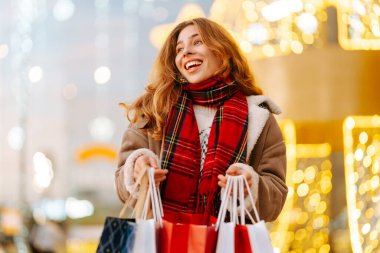 Alışveriş torbaları taşıyan neşeli bir kadın modern bir alışveriş merkezinin arka planında duruyor. Kadınlar açık havada alışveriş yaptıktan sonra şenlik havasına girerler. Konseptler: indirim, kutlama, alışveriş.