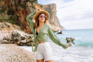 Mutlu kadın kayalık sahilde hasır şapka takıp güneşli bir günde deniz esintisinin tadını çıkarıyor. Güzel bir kadın deniz kıyısında rahatlıyor, özgürlüğü ve rüzgarı saçlarında hissediyor. Rahatlama ve keyif kavramı.