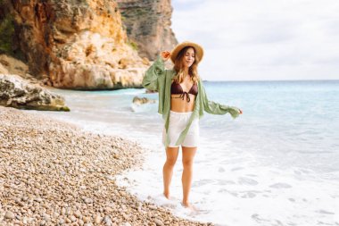 Mutlu kadın kayalık sahilde hasır şapka takıp güneşli bir günde deniz esintisinin tadını çıkarıyor. Güzel bir kadın deniz kıyısında rahatlıyor, özgürlüğü ve rüzgarı saçlarında hissediyor. Rahatlama ve keyif kavramı.