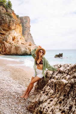 Mutlu kadın kayalık sahilde hasır şapka takıp güneşli bir günde deniz esintisinin tadını çıkarıyor. Güzel bir kadın deniz kıyısında rahatlıyor, özgürlüğü ve rüzgarı saçlarında hissediyor. Rahatlama ve keyif kavramı.