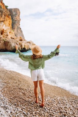 Mutlu kadın kayalık sahilde hasır şapka takıp güneşli bir günde deniz esintisinin tadını çıkarıyor. Güzel bir kadın deniz kıyısında rahatlıyor, özgürlüğü ve rüzgarı saçlarında hissediyor. Rahatlama ve keyif kavramı.