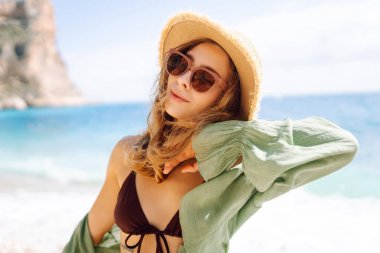 Mutlu kadın kayalık sahilde hasır şapka takıp güneşli bir günde deniz esintisinin tadını çıkarıyor. Güzel bir kadın deniz kıyısında rahatlıyor, özgürlüğü ve rüzgarı saçlarında hissediyor. Rahatlama ve keyif kavramı.