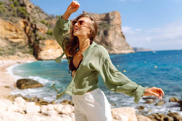 Mutlu kadın kayalık sahilde hasır şapka takıp güneşli bir günde deniz esintisinin tadını çıkarıyor. Güzel bir kadın deniz kıyısında rahatlıyor, özgürlüğü ve rüzgarı saçlarında hissediyor. Rahatlama ve keyif kavramı.