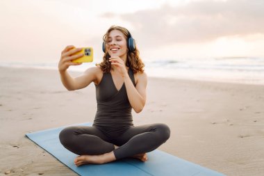 Spor kıyafetli, elinde telefonla gülümseyen kadın, denize bakan yoga minderinin üzerinde duruyor. Kulaklıkları olan atletik bir kadın. Plajda esneme hareketleri, egzersizler ve bloglar yapıyor. Konseptler: blog yazma, spor, rahatlama.