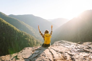 Sarı sırt çantalı bir bayan yürüyüşçü bir dağın kayalık kenarında duruyor. Genç bir kadın gün batımında dağ manzarasından zevk alır. Arkadan bak. Yürüyüş, rahatlama, doğa kavramları..