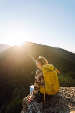 Sarı sırt çantalı bir bayan yürüyüşçü bir dağın kayalık kenarında duruyor. Genç bir kadın gün batımında dağ manzarasından zevk alır. Arkadan bak. Yürüyüş, rahatlama, doğa kavramları..