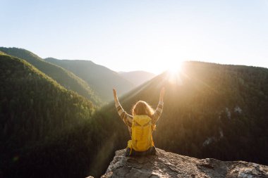 Sarı sırt çantalı bir bayan yürüyüşçü bir dağın kayalık kenarında duruyor. Genç bir kadın gün batımında dağ manzarasından zevk alır. Arkadan bak. Yürüyüş, rahatlama, doğa kavramları..