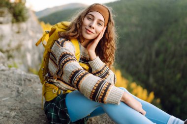 Sarı sırt çantalı bir bayan yürüyüşçü bir dağın kayalık kenarında duruyor. Genç bir kadın gün batımında dağ manzarasından zevk alır. Arkadan bak. Yürüyüş, rahatlama, doğa kavramları..