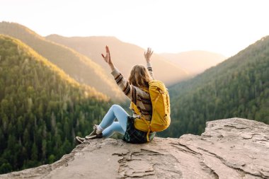 Sarı sırt çantalı bir bayan yürüyüşçü bir dağın kayalık kenarında duruyor. Genç bir kadın gün batımında dağ manzarasından zevk alır. Arkadan bak. Yürüyüş, rahatlama, doğa kavramları..