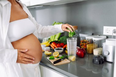 Hamile bir kadın modern bir mutfakta sağlıklı bir içecek hazırlar. Bebek bekleyen genç bir kadın sıcak bir odada güneşli bir günün tadını çıkarıyor. Gıda, sağlık ve beklenti kavramları.