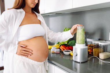 Hamile bir kadın modern bir mutfakta sağlıklı bir içecek hazırlar. Bebek bekleyen genç bir kadın sıcak bir odada güneşli bir günün tadını çıkarıyor. Gıda, sağlık ve beklenti kavramları.