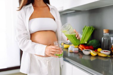 Hamile bir kadın modern bir mutfakta sağlıklı bir içecek hazırlar. Bebek bekleyen genç bir kadın sıcak bir odada güneşli bir günün tadını çıkarıyor. Gıda, sağlık ve beklenti kavramları.