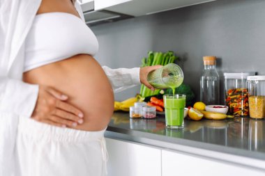 Hamile bir kadın modern bir mutfakta sağlıklı bir içecek hazırlar. Bebek bekleyen genç bir kadın sıcak bir odada güneşli bir günün tadını çıkarıyor. Gıda, sağlık ve beklenti kavramları.