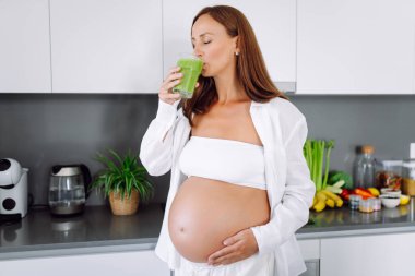 Hamile bir kadın modern bir mutfakta sağlıklı bir içecek hazırlar. Bebek bekleyen genç bir kadın sıcak bir odada güneşli bir günün tadını çıkarıyor. Gıda, sağlık ve beklenti kavramları.