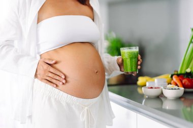 Hamile bir kadın modern bir mutfakta sağlıklı bir içecek hazırlar. Bebek bekleyen genç bir kadın sıcak bir odada güneşli bir günün tadını çıkarıyor. Gıda, sağlık ve beklenti kavramları.