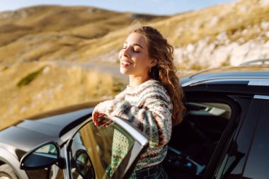 Dağlık bir bölgede siyah arabanın yanında duran şapkalı bir gezgin. Yolculuktaki güzel kadın kendini özgür hissediyor ve dağ manzarasından hoşlanıyor. Seyahat, boş zaman ve ulaşım kavramları.