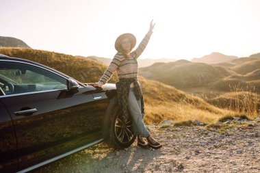 Dağlık bir bölgede siyah arabanın yanında duran şapkalı bir gezgin. Yolculuktaki güzel kadın kendini özgür hissediyor ve dağ manzarasından hoşlanıyor. Seyahat, boş zaman ve ulaşım kavramları.