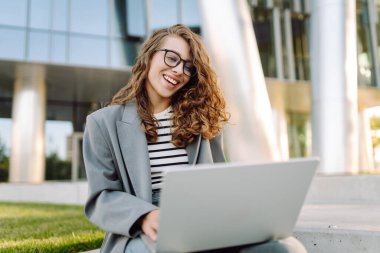 Tarz sahibi bir kadın dizüstü bilgisayarda çalışırken arka planda modern binaların olduğu merdivenlerde oturuyor. Gözlüklü güzel kadın dışarıda dizüstü bilgisayar kullanıyor. Konsept: uzak çalışma, tarz ve moda.