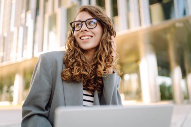 Tarz sahibi bir kadın dizüstü bilgisayarda çalışırken arka planda modern binaların olduğu merdivenlerde oturuyor. Gözlüklü güzel kadın dışarıda dizüstü bilgisayar kullanıyor. Konsept: uzak çalışma, tarz ve moda.