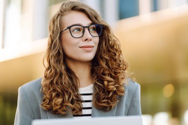 Tarz sahibi bir kadın dizüstü bilgisayarda çalışırken arka planda modern binaların olduğu merdivenlerde oturuyor. Gözlüklü güzel kadın dışarıda dizüstü bilgisayar kullanıyor. Konsept: uzak çalışma, tarz ve moda.