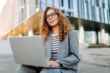 Tarz sahibi bir kadın dizüstü bilgisayarda çalışırken arka planda modern binaların olduğu merdivenlerde oturuyor. Gözlüklü güzel kadın dışarıda dizüstü bilgisayar kullanıyor. Konsept: uzak çalışma, tarz ve moda.