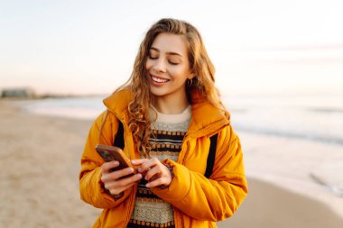 Telefonu olan genç bir kadın gün batımında kumlu bir sahilde duruyor. Güzel bir kadın deniz manzarasını ve blogları sever. Yürüme, doğa ve teknoloji kavramı.