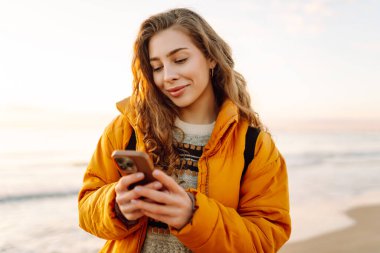 Telefonu olan genç bir kadın gün batımında kumlu bir sahilde duruyor. Güzel bir kadın deniz manzarasını ve blogları sever. Yürüme, doğa ve teknoloji kavramı.