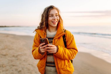 A young woman with a phone stands on a sandy beach at sunset. A beautiful woman enjoys the seascape and blogs outdoors. Concept of walking, nature, and technology.