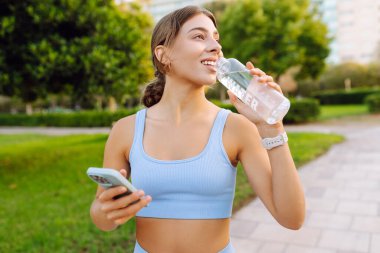 Spor giyimli neşeli bir kadın güneşli bir parkta elinde bir şişe su ve telefonla dikiliyor. Güzel bir kadın idmandan sonra su içer ve mesajlaşır. Konsept: Spor, eğlence. Etkin yaşam biçimi.