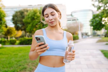 Spor giyimli neşeli bir kadın güneşli bir parkta elinde bir şişe su ve telefonla dikiliyor. Güzel bir kadın idmandan sonra su içer ve mesajlaşır. Konsept: Spor, eğlence. Etkin yaşam biçimi.