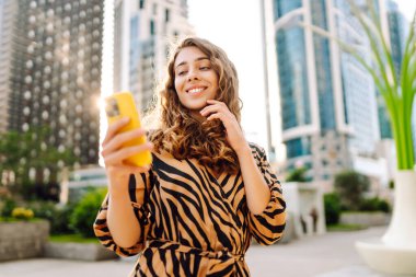 Güneşli bir caddede elinde telefonla duran gülümseyen bir kadının portresi. Güzel bir kadın blogu, metropol manzaralı bir gezintiden keyif alıyor. Blogculuk ve turizm kavramları.