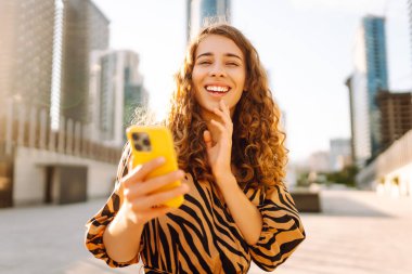 Güneşli bir caddede elinde telefonla duran gülümseyen bir kadının portresi. Güzel bir kadın blogu, metropol manzaralı bir gezintiden keyif alıyor. Blogculuk ve turizm kavramları.