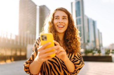 Güneşli bir caddede elinde telefonla duran gülümseyen bir kadının portresi. Güzel bir kadın blogu, metropol manzaralı bir gezintiden keyif alıyor. Blogculuk ve turizm kavramları.